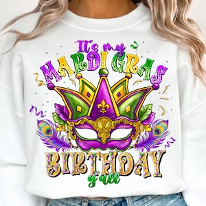 May include: White sweatshirt with a colorful Mardi Gras design. The graphic includes a purple and gold mask, a crown, and the text "It's my Mardi Gras Birthday y'all" in various colors. Features peacock feathers and confetti.