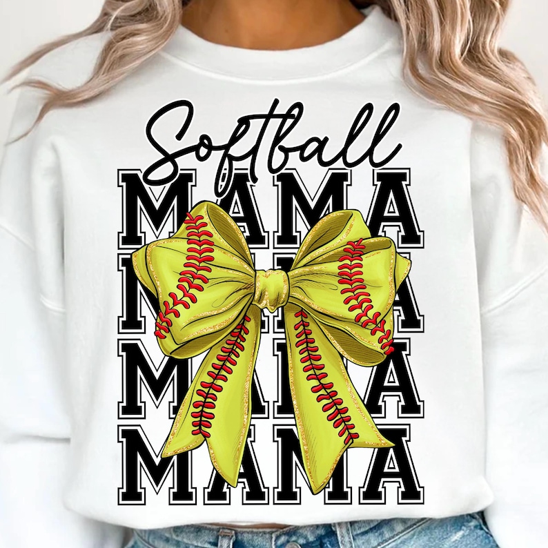 May include: White sweatshirt with the words "Softball Mama" in black script and block letters. A large yellow bow with red stitching, resembling a softball, is the central design element.
