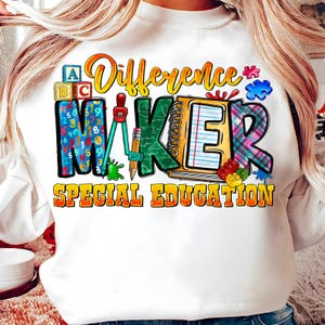 Pode incluir: Camisola branca com as palavras "Difference Maker" e "Special Education" num design colorido. O design inclui blocos ABC, um caderno, um compasso e peças de quebra-cabeça.
