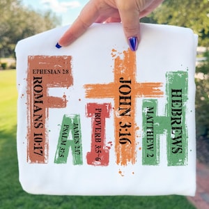 May include: White sweatshirt with the word "FAITH" in colorful, textured lettering. The letters are composed of various Bible verses, including Romans 10:17, John 3:16, and Hebrews. The design has a hand-painted aesthetic.