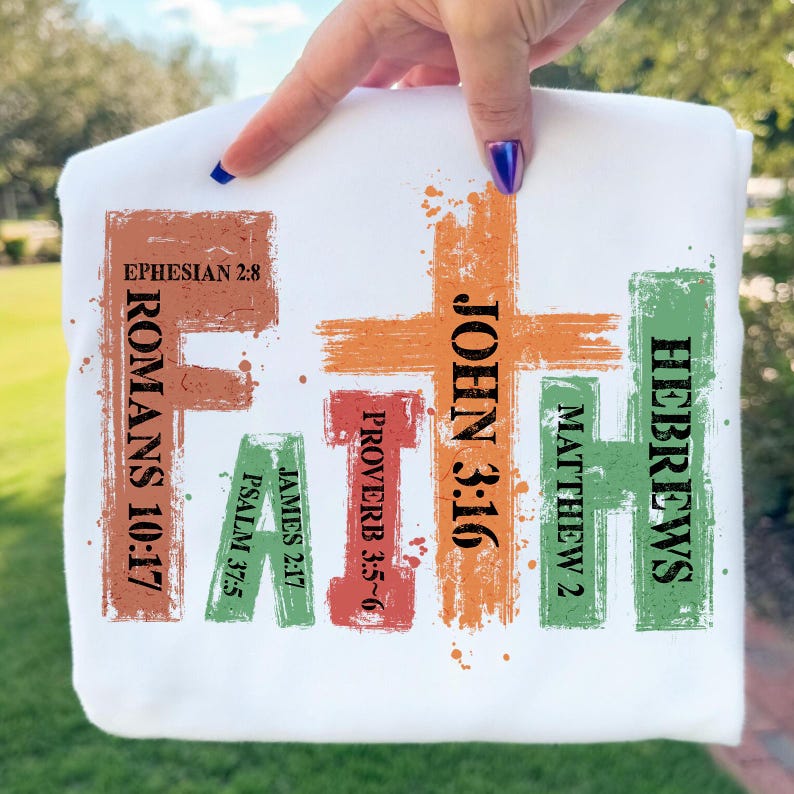 May include: White sweatshirt with the word "FAITH" in colorful, textured lettering. The letters are composed of various Bible verses, including Romans 10:17, John 3:16, and Hebrews. The design has a hand-painted aesthetic.