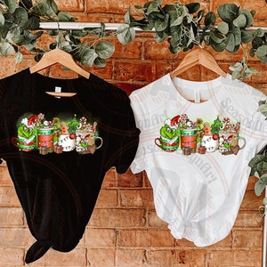 May include: Black and white t-shirts with a Christmas-themed graphic. The design features mugs with festive characters, candy canes, and gingerbread men. The shirts are hanging on wooden hangers against a brick wall.