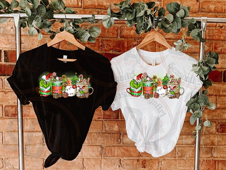 May include: Black and white t-shirts with a Christmas-themed graphic. The design features mugs with festive characters, candy canes, and gingerbread men. The shirts are hanging on wooden hangers against a brick wall.