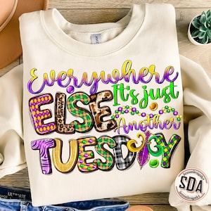 May include: A white sweatshirt with the text "Everywhere Else It's Just Another Tuesday" in purple, green, and gold letters. The letters are decorated with Mardi Gras beads, leopard print, and plaid patterns.
