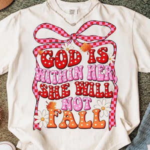 May include: A white short-sleeve t-shirt featuring a large graphic of a pink and red checkered bow. The text "GOD IS WITHIN HER, SHE WILL NOT FALL" is printed in red, pink, and orange bubble letters with white polka dots, accented by daisies and leaves.