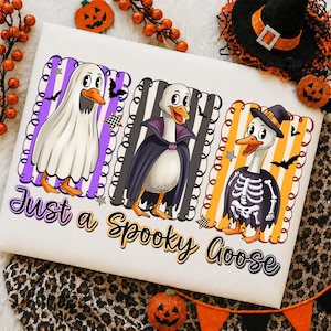 May include: A white t-shirt featuring three cartoon geese dressed in Halloween costumes: a ghost, a vampire, and a skeleton. The geese are positioned over purple, gray, and orange striped backgrounds. The text reads Just a Spooky Goose.