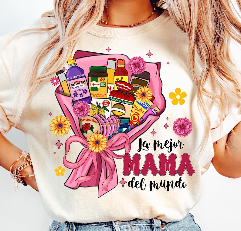 May include: A white t-shirt with a pink and yellow floral design. The design features a bouquet of various Mexican grocery items, including a bottle of Fabuloso, a jar of Mama brand salsa, and a box of La Banderita tortillas. The text "La mejor MAMA del mundo" is printed below the bouquet.