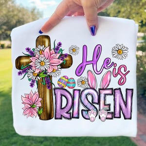 May include: White sweatshirt featuring a wooden cross adorned with pink flowers, lavender, and daisies. The text "He is Risen" is in purple and pink, with bunny ears and Easter eggs. A festive design for Easter.