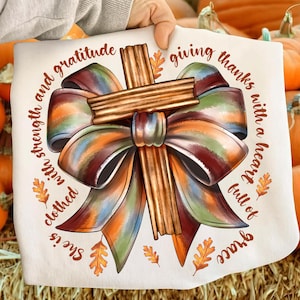 May include: A cream-colored decorative pillow featuring a cross made of cinnamon sticks and a large, colorful bow. The text around the bow reads "She is clothed with strength and gratitude, giving thanks with a heart full of grace."