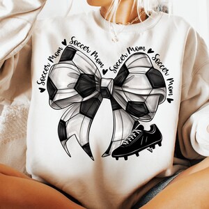 May include: Cream-colored sweatshirt featuring a soccer ball bow design with a black soccer cleat. The words "Soccer Mom" are printed around the bow. The bow is black and white.