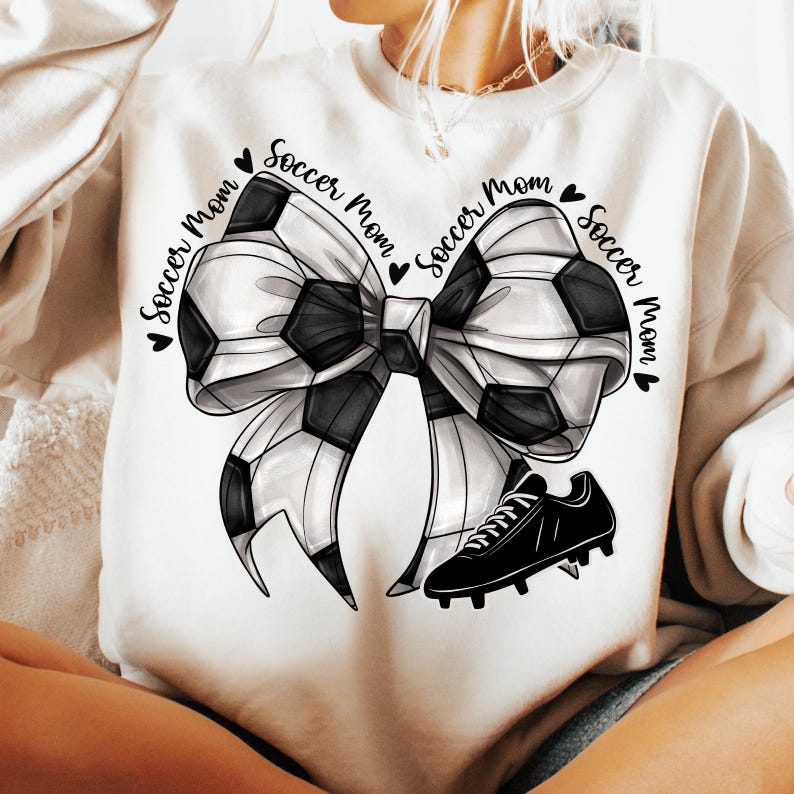 May include: Cream-colored sweatshirt featuring a soccer ball bow design with a black soccer cleat. The words "Soccer Mom" are printed around the bow. The bow is black and white.