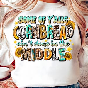Puede incluir: Sudadera blanca con la frase "SOME OF Y'ALLS CORNBREAD ain't done in the MIDDLE". El texto es multicolor con diseños de girasoles y estrellas. Una prenda superior informal y divertida.