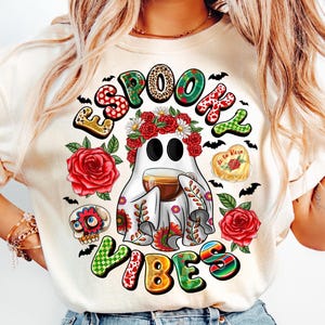 May include: Cream-colored t-shirt with a colorful Halloween-themed graphic. The design features a ghost wearing a floral crown and holding a cup, surrounded by roses, bats, and the words "SPOOKY VIBES".