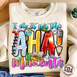Puede incluir: Una sudadera blanca con un diseño gráfico colorido que dice "I do it for the AHA! moments". El gráfico incluye un globo terráqueo, una regla, un lápiz, un libro, una manzana roja y un clip.