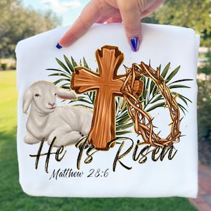 May include: White sweatshirt featuring a lamb, wooden cross, and crown of thorns. The words "He Is Risen" and "Matthew 28:6" are printed in black script. Green leaves and gold accents complete the design.