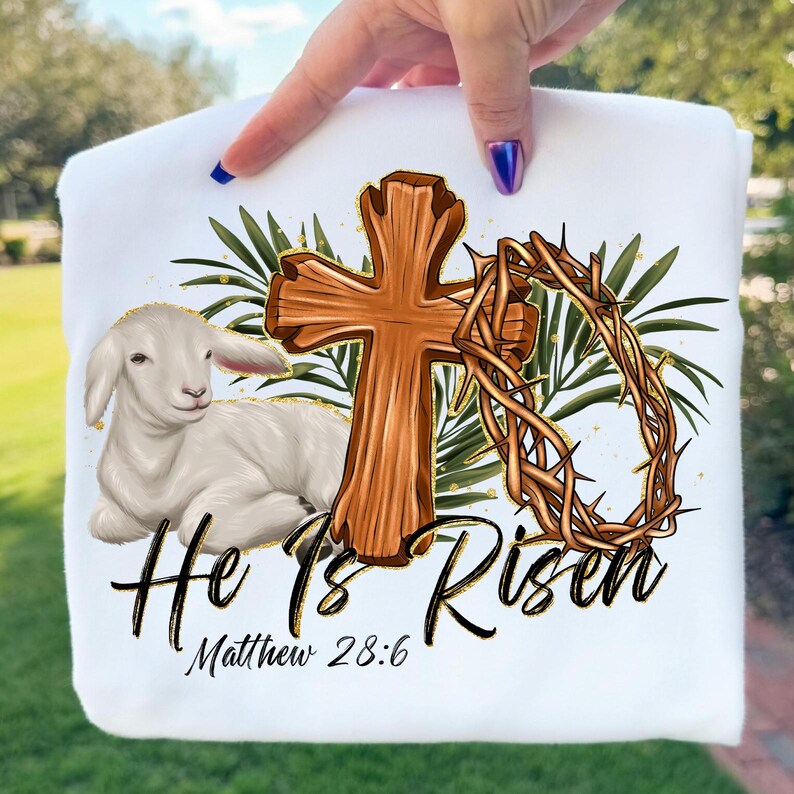 May include: White sweatshirt featuring a lamb, wooden cross, and crown of thorns. The words "He Is Risen" and "Matthew 28:6" are printed in black script. Green leaves and gold accents complete the design.