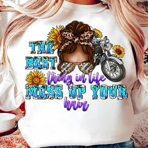 May include: A white sweatshirt featuring a graphic of a person with a messy bun, leopard print headband, and sunglasses, alongside a motorcycle and sunflowers. The text reads: THE BEST thing in life MESS UP YOUR hair in blue and purple.