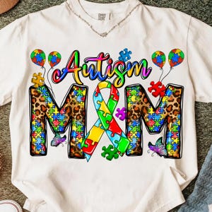 May include: White t-shirt with the words "Autism M&M" in colorful lettering and puzzle piece designs. The letters "M&M" are filled with a leopard print pattern. The design includes puzzle piece balloons and butterflies.