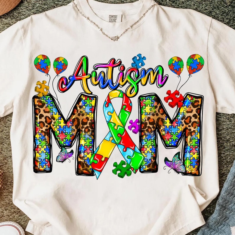 May include: White t-shirt with the words "Autism M&M" in colorful lettering and puzzle piece designs. The letters "M&M" are filled with a leopard print pattern. The design includes puzzle piece balloons and butterflies.
