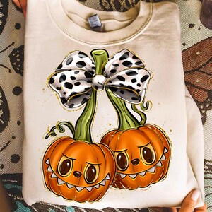Könnte beinhalten: Cremefarbenes Sweatshirt mit einem Halloween-Motiv. Die Grafik zeigt zwei orangefarbene Kürbisse mit lächelnden Gesichtern, verbunden durch grüne Stiele und eine große Schleife mit schwarzen Punkten. Goldakzente und Details werten das Design auf.