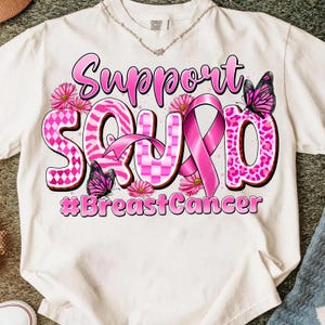 May include: White t-shirt with a pink and black graphic that says "Support Squad" with a pink ribbon and butterflies. The text "#BreastCancer" is below the main text. The shirt has a crew neck and short sleeves.