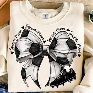 May include: Cream-colored sweatshirt featuring a soccer ball bow design with the words "Soccer Mom" in a heart-shaped design. A black soccer cleat is positioned below the bow. The design is in black and white.