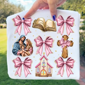 May include: White fabric featuring religious illustrations and pink bows with gold glitter accents. The images include a depiction of the Holy Family, a church, a cross, a rosary, and an open book.