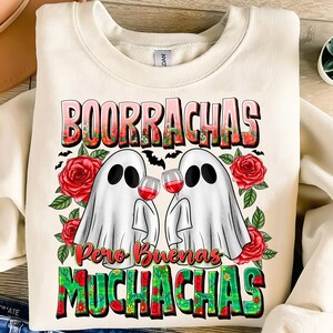 May include: Cream-colored sweatshirt with a graphic featuring two white ghost figures holding wine glasses. The text "BOORRACHAS Pero Buenas MUCHACHAS" is displayed in a colorful, floral design, with red roses and bats.