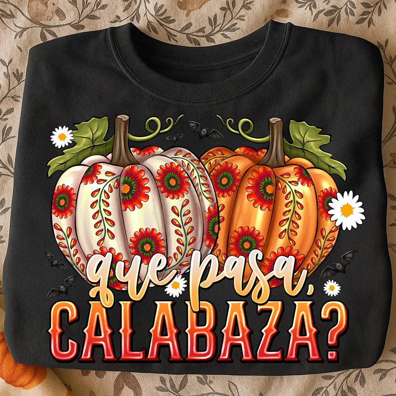 May include: A black sweatshirt featuring a graphic of two pumpkins, one white and one orange, decorated with red and green floral patterns. The design includes small white daisies, black bats, and the text "que pasa, CALABAZA?" in a stylized font.