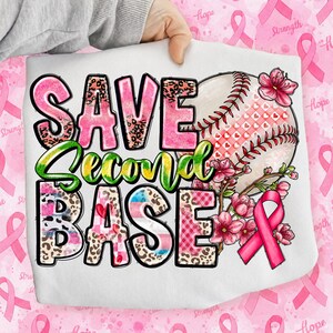 May include: A white t-shirt featuring the text SAVE SECOND BASE in pink, leopard print, and green gradient lettering. A baseball graphic with a red stitched pattern, pink flowers, and a pink awareness ribbon are printed on the right side.
