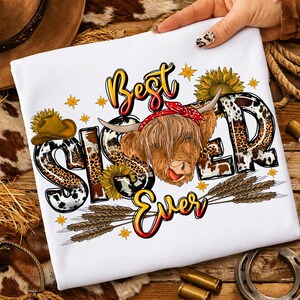 May include: White t-shirt with the words "Best Sister Ever" in a colorful, stylized font. The design includes a Highland cow wearing a red bandana, sunflowers, stars, and wheat stalks. The word "Sister" has a leopard print pattern.