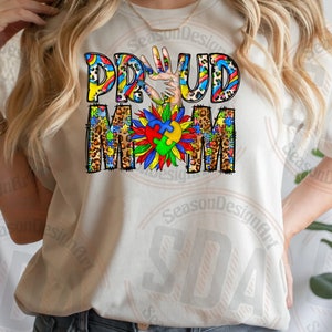K&ouml;nnte beinhalten: Wei&szlig;es T-Shirt mit den Worten "Proud Mom" in bunten Buchstaben mit einem Puzzle-Motiv und einem Handabdruck. Die Buchstaben sind mit Leopardenmuster verziert.