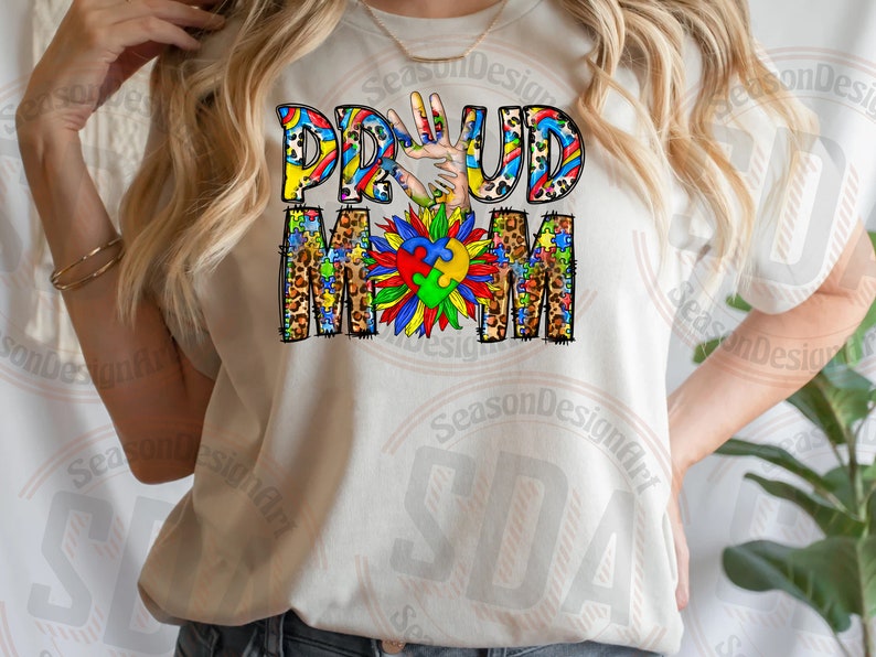 K&ouml;nnte beinhalten: Wei&szlig;es T-Shirt mit den Worten "Proud Mom" in bunten Buchstaben mit einem Puzzle-Motiv und einem Handabdruck. Die Buchstaben sind mit Leopardenmuster verziert.