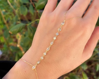May include: A delicate gold hand chain bracelet featuring a series of clear, sparkling gemstones. The bracelet connects to a ring and drapes elegantly across the back of the hand. The gemstones are set in a gold chain.