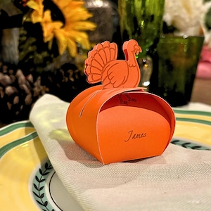 Puede incluir: Caja de regalo de papel naranja en forma de pavo con el nombre "James" escrito en la parte delantera. La caja está sobre una servilleta de papel blanca y un plato amarillo y verde.