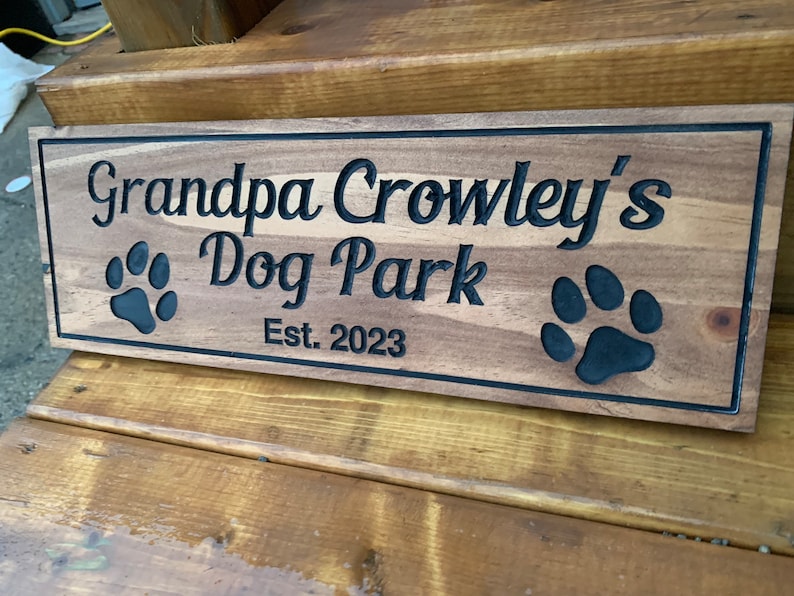 May include: A wooden sign with the text "Grandpa Crowley's Dog Park Est. 2023" and two black paw prints.