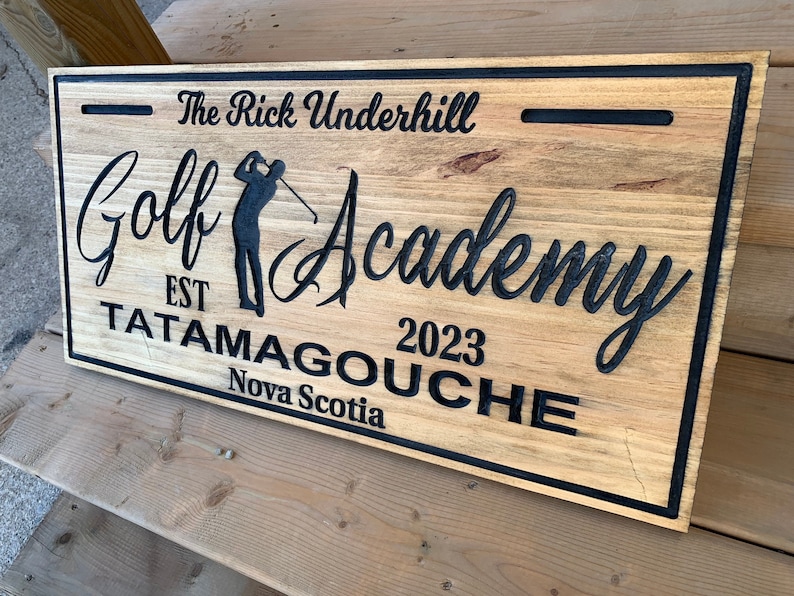 May include: A wooden sign with black lettering that reads "The Rick Underhill Golf Academy Est 2023 Tatamagouche Nova Scotia". The sign features a silhouette of a golfer swinging a club.