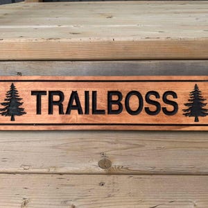 Peut inclure: Une pancarte en bois avec le texte "TRAILBOSS" en lettres noires. La pancarte est décorée de deux sapins noirs en silhouette de chaque côté du texte.
