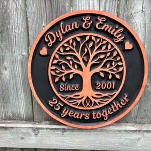 May include: A round wooden sign with a black background and an orange border. The sign features a tree of life design in the center, with the names "Dylan & Emily" and the text "25 years together" and "Since 2001".