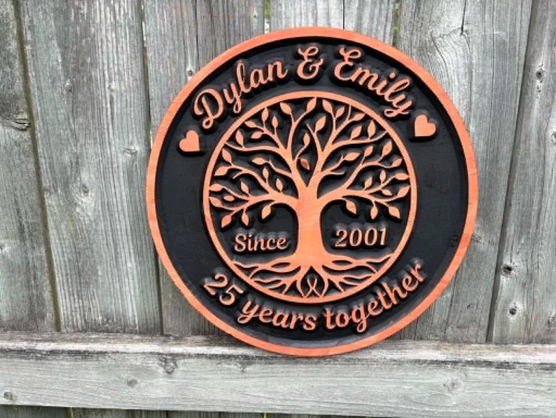 May include: A round wooden sign with a black background and an orange border. The sign features a tree of life design in the center, with the names "Dylan & Emily" and the text "25 years together" and "Since 2001".