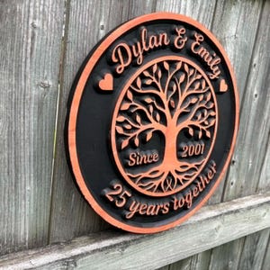 May include: A round, wooden plaque with a black border and an orange interior. The plaque features the names "Dylan & Emily" and the text "25 years together" with a tree of life design. The year "2001" is also included.