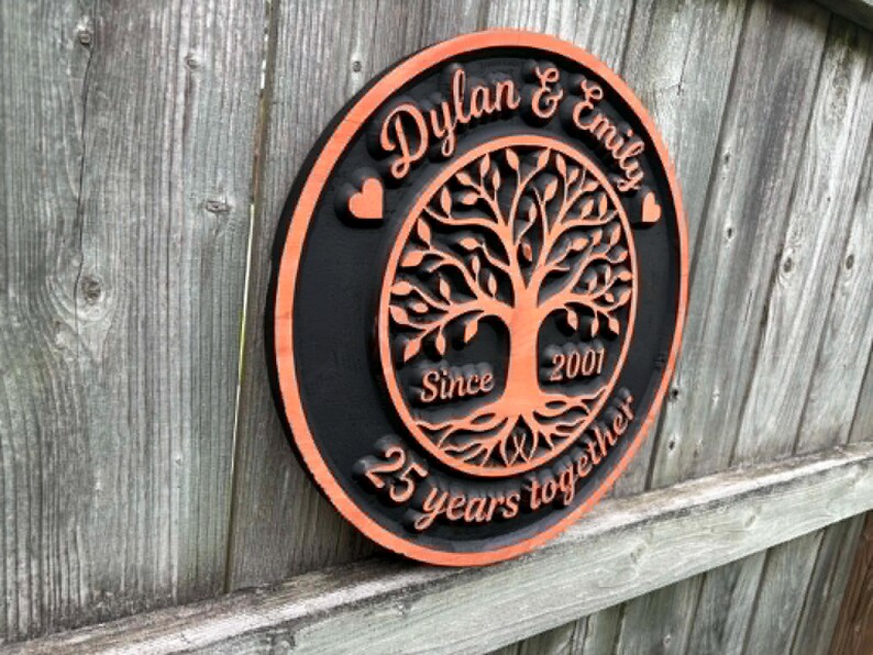 May include: A round, wooden plaque with a black border and an orange interior. The plaque features the names "Dylan & Emily" and the text "25 years together" with a tree of life design. The year "2001" is also included.