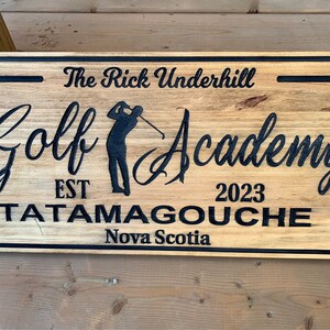 May include: A wooden sign with black lettering that reads "The Rick Underhill Golf Academy Est 2023 Tatamagouche Nova Scotia". The sign features a silhouette of a golfer swinging a club.