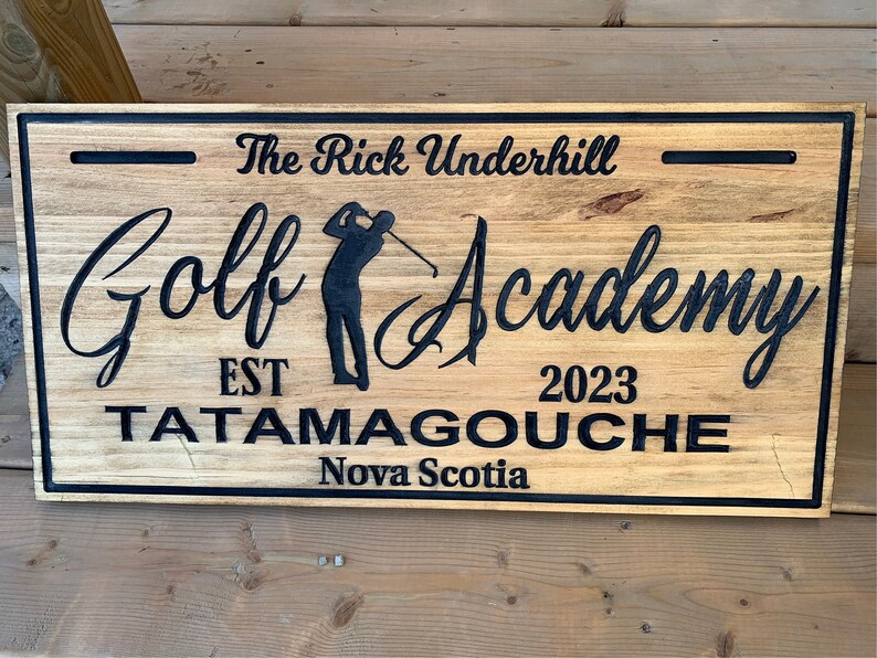 May include: A wooden sign with black lettering that reads "The Rick Underhill Golf Academy Est 2023 Tatamagouche Nova Scotia". The sign features a silhouette of a golfer swinging a club.