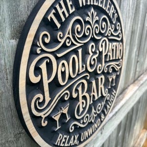 Puede incluir: Un letrero redondo de madera con borde negro y letras de madera clara. El letrero dice "The Wheeler's Pool & Patio Bar" con adornos decorativos. Las palabras "Relax, Unwind & Laugh" están en la parte inferior. El letrero está montado en una valla de madera.
