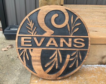 Puede incluir: Un letrero redondo de madera con fondo negro y un patrón de veta de madera marrón. El letrero tiene un diseño tallado de un remolino estilizado y hojas. El nombre "EVANS" está tallado en el centro del letrero.