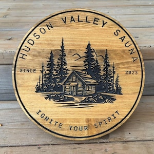 May include: A round wooden sign with the text "Hudson Valley Sauna" and "Ignite Your Spirit" around the edge. The sign also features a cabin in the woods with the text "Since 2023".