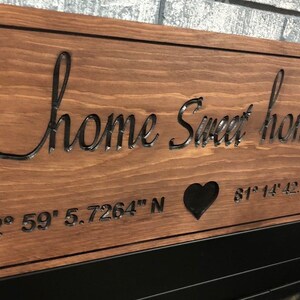 Peut inclure: Panneau en bois marron avec l'inscription "home Sweet home" en lettres cursives. Le panneau comporte un c&oelig;ur et des coordonn&eacute;es g&eacute;ographiques : 42&deg; 59' 5.7264" N et 81&deg; 14' 42.9972" W. Bordure noire.