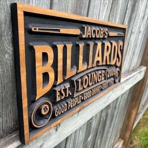 May include: A rectangular wooden sign with a black background and a brown frame. The sign reads "Jacob's Billiards Lounge" with the establishment year "2026" and the phrase "Good People Good Drinks Great Times."