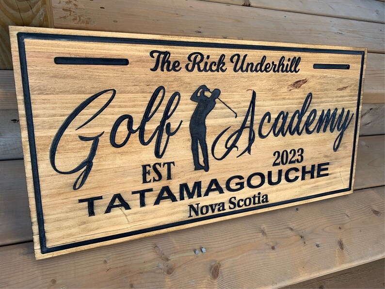 May include: A wooden sign with black lettering that reads "The Rick Underhill Golf Academy Est 2023 Tatamagouche Nova Scotia". The sign features a silhouette of a golfer swinging a club.